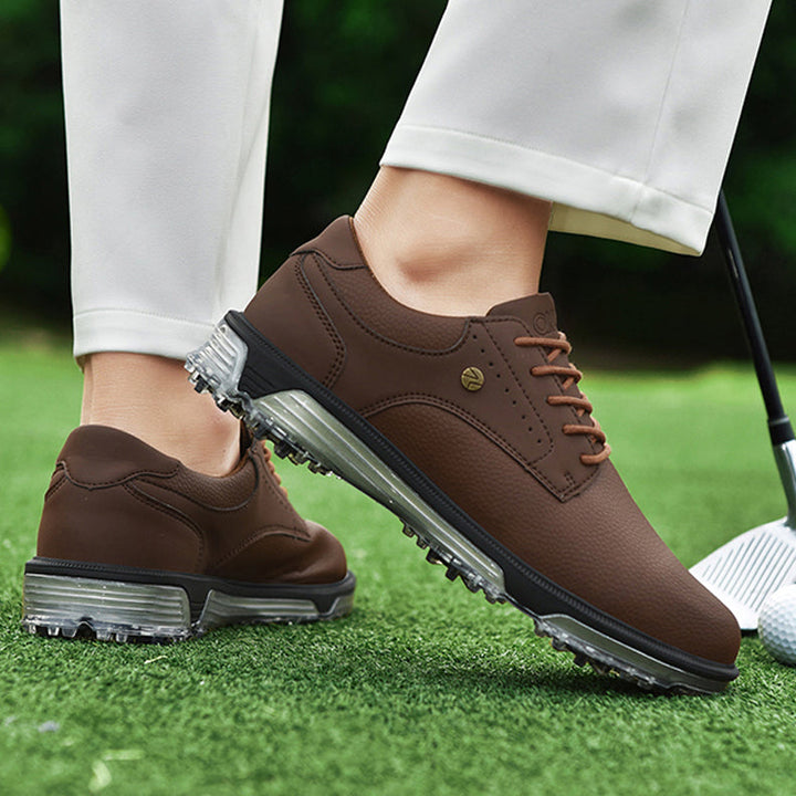 A person wearing brown, genuine leather golf shoes with clear, spiked soles stands on green grass, holding a golf club. The advanced grip technology ensures stability. The person is dressed in light-colored pants against a blurred greenery background.