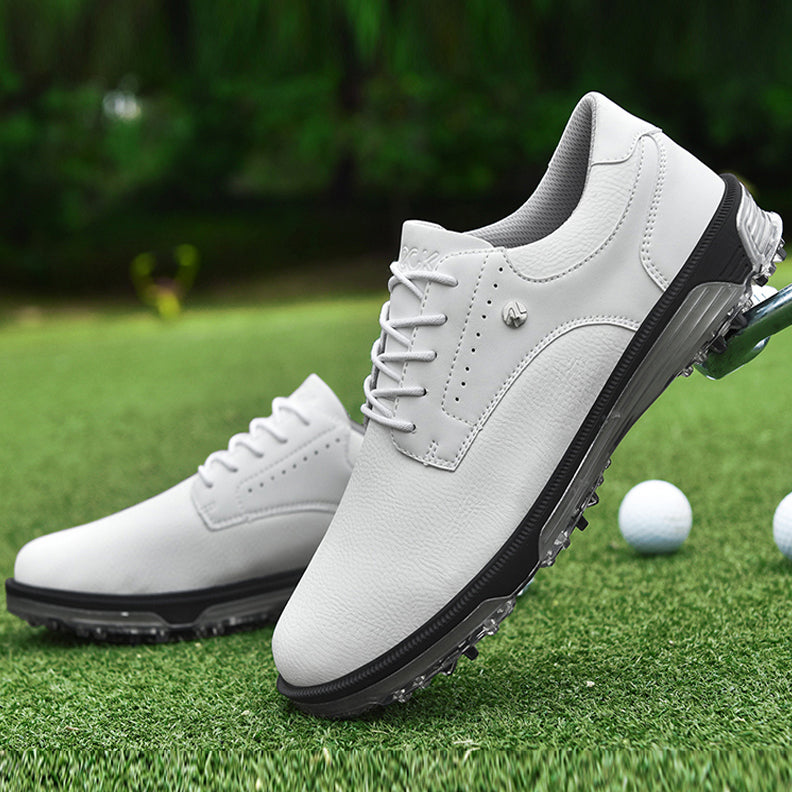 A pair of genuine leather golf shoes with advanced grip technology and black soles is displayed on green grass beside three white golf balls, set against a blurry outdoor background of lush greenery.