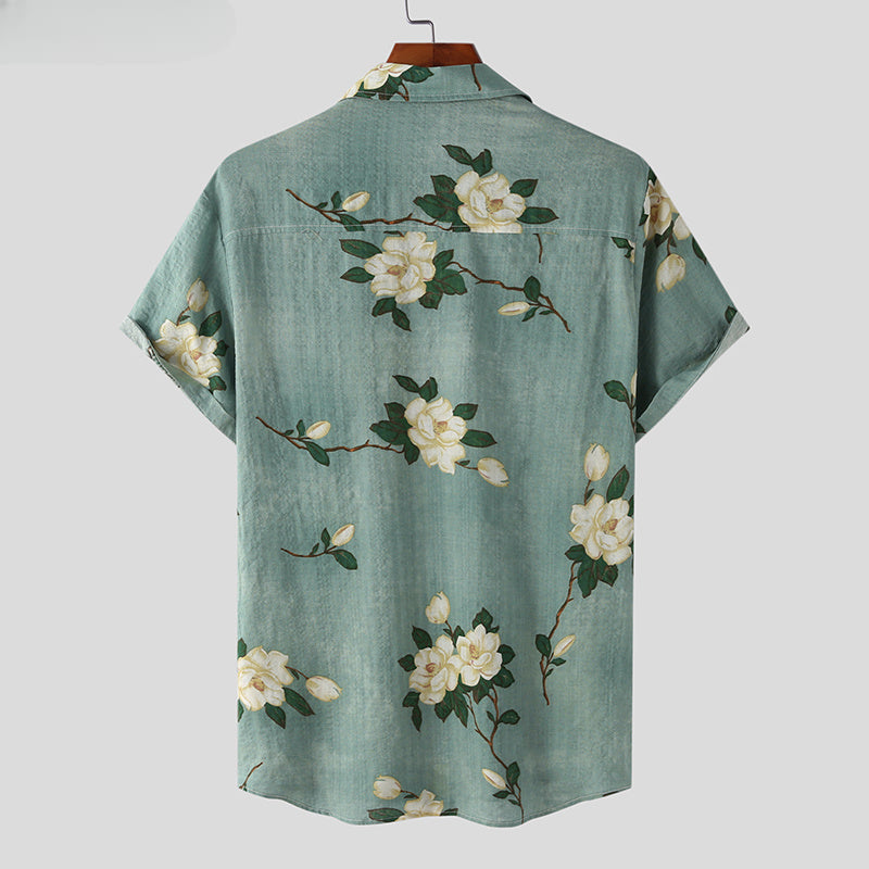 A light blue short-sleeve button-up Mens Hawaiian Shirt with white magnolia flower prints and green leaf details, shown from the back on a hanger against a plain background.