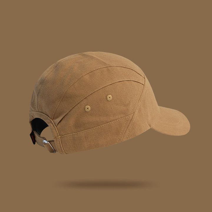 A tan unisex cap made of textured fabric is shown on a solid brown background. The Japanese-style peaked cap is viewed from the back and side, featuring an adjustable strap with a metal buckle. A faint shadow appears below the cap.