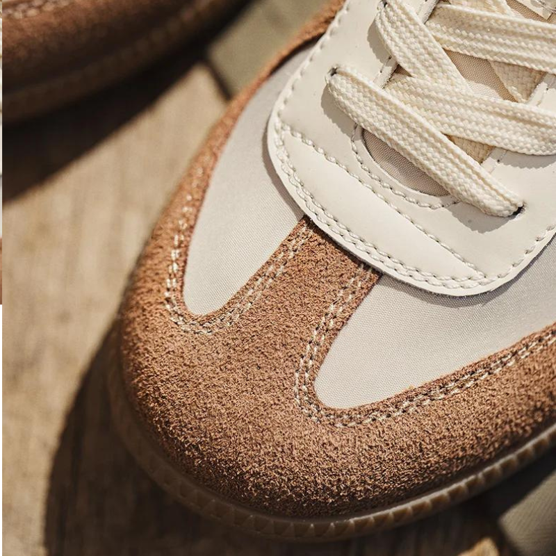Close-up of a sneaker with premium cow suede, white leather, beige fabric details on the toe, and cream-colored laces—highlighting lightweight breathable sneakers on a wooden surface.