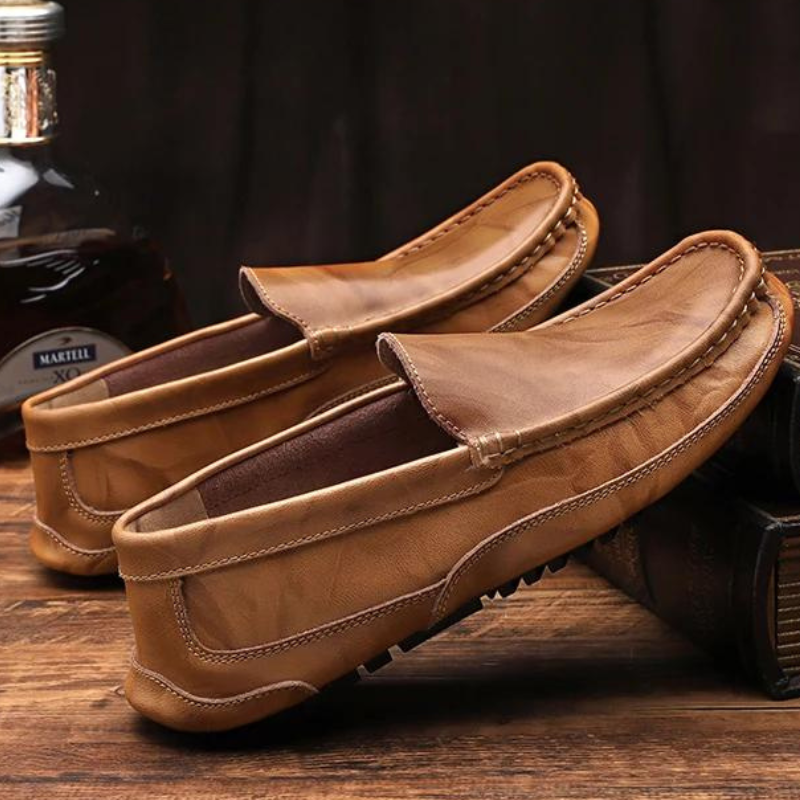 A pair of genuine leather loafers is displayed on a wooden surface, with one shoe resting atop a closed book and a bottle in the background. The hand-stitched moccasin design adds an artisanal touch to these classic shoes.