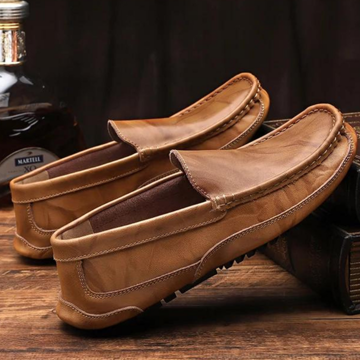 A pair of genuine leather loafers is displayed on a wooden surface, with one shoe resting atop a closed book and a bottle in the background. The hand-stitched moccasin design adds an artisanal touch to these classic shoes.