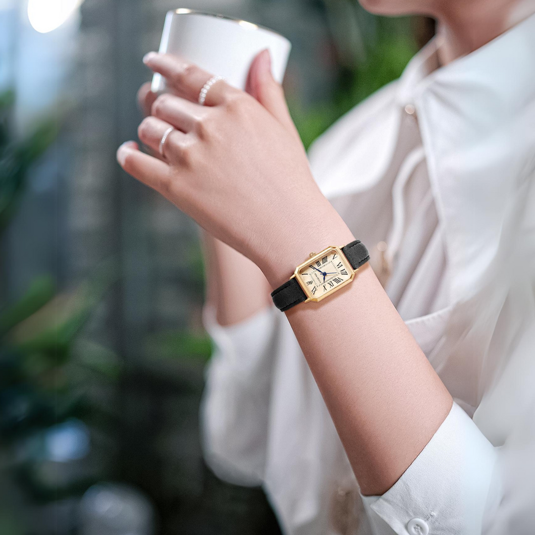 A person wearing a gold rectangular face watch with a genuine leather strap holds a white cup. They are dressed in a white blouse, and greenery is visible in the blurred background.