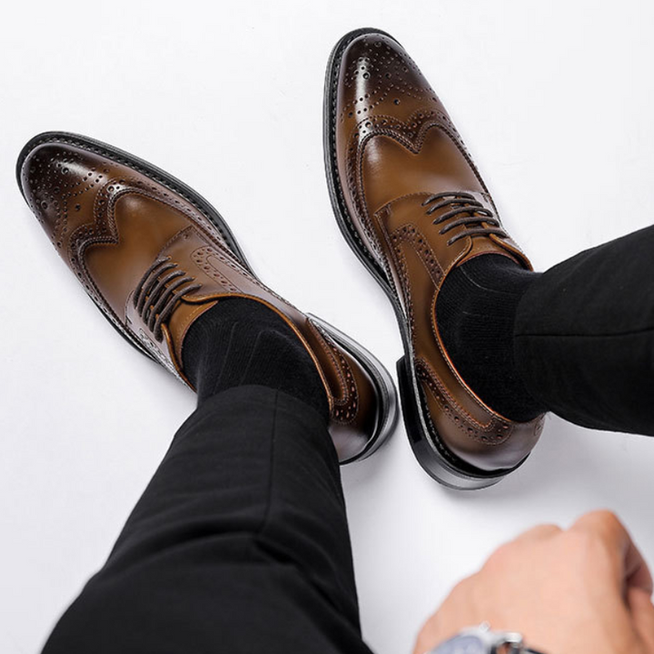 A person wearing black pants and black socks with brown premium leather brogue dress shoes featuring decorative perforations and a slip-resistant rubber sole, shown from above on a white background.