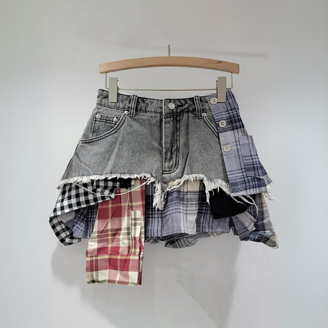 A patchwork skirt with a frayed denim top and plaid, checkered panels in red, black, white, and blue hangs on a wooden hanger against a plain white background—perfect for lovers of eclectic fashion.