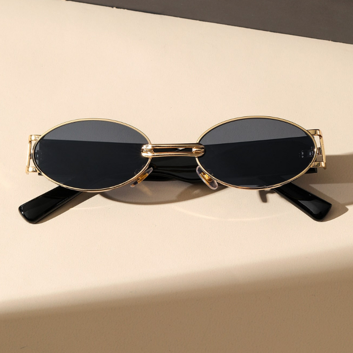 A pair of retro oval sunglasses with UV400 protection, featuring dark lenses and a durable metal frame in gold, rest on a light beige surface with the black arms crossed underneath.