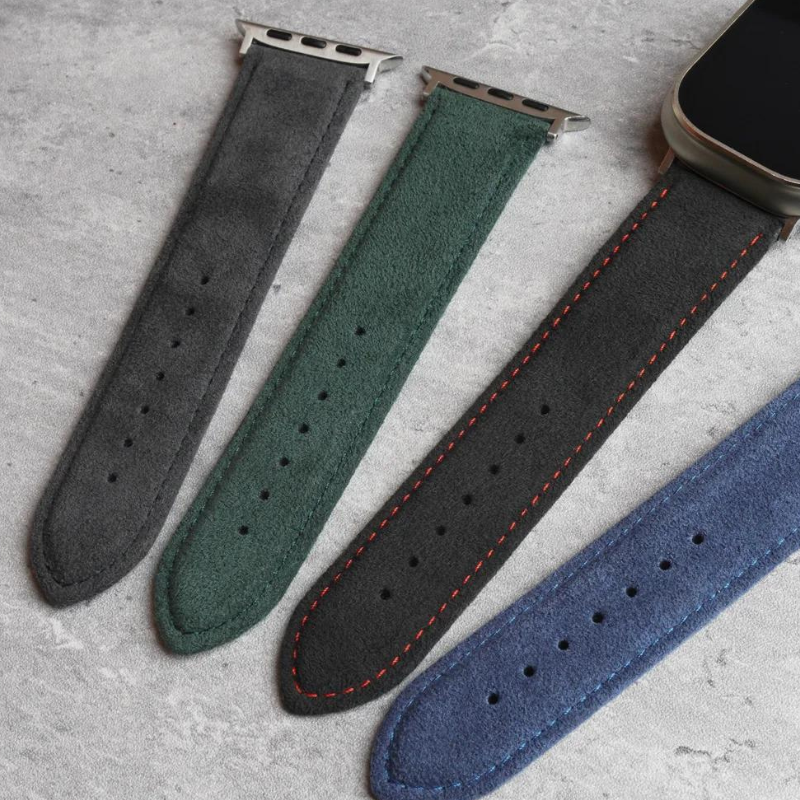 Four suede watch bands in grey, green, black with orange stitching, and blue are laid out side by side on a grey textured surface; one band is attached to a smartwatch and features a precision fit with a stainless steel buckle.