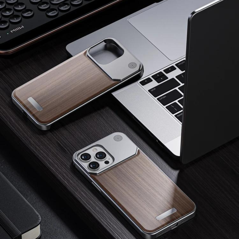 A smartphone in a stylish phone case lies on a dark wooden desk near a matching wood phone case, a laptop, a keyboard, and a notebook.