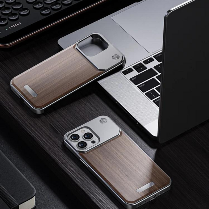 A smartphone in a stylish phone case lies on a dark wooden desk near a matching wood phone case, a laptop, a keyboard, and a notebook.