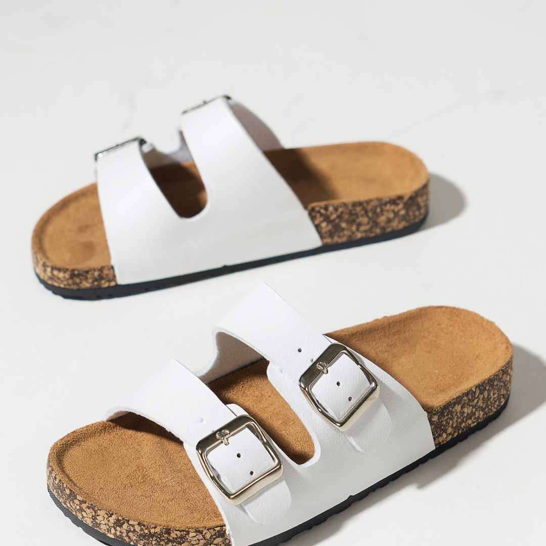A pair of white double-buckle sandals with premium leather straps and ergonomic cork footbed, displayed on a light surface.