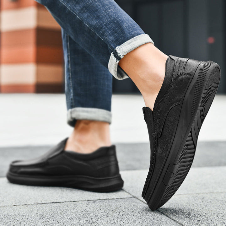 Close-up of a person wearing black Slip-On Men’s Cowhide Leather Shoes with a Soft Sole and rolled-up blue jeans, walking on a paved outdoor surface with a blurred building in the background.