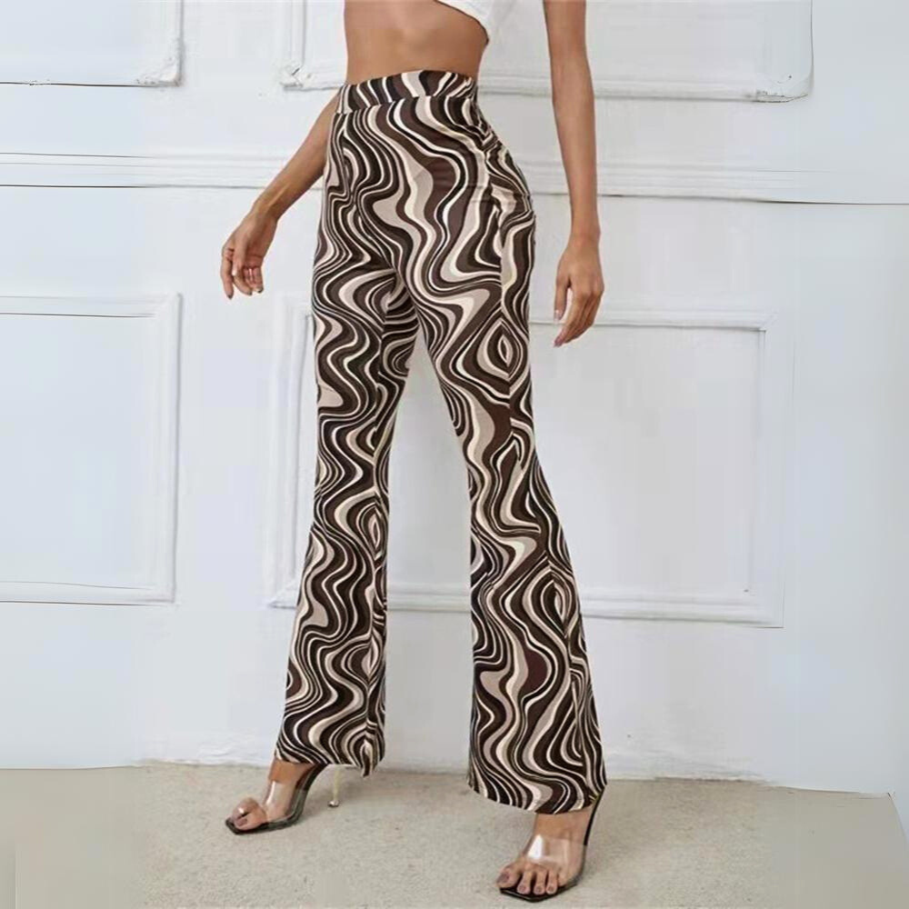 A person wearing retro-inspired, brown and beige swirl-patterned flared pants and a white crop top stands indoors, with one arm relaxed by their side and clear-heeled sandals completing the look.