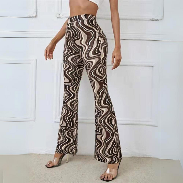 A person wearing retro-inspired, brown and beige swirl-patterned flared pants and a white crop top stands indoors, with one arm relaxed by their side and clear-heeled sandals completing the look.
