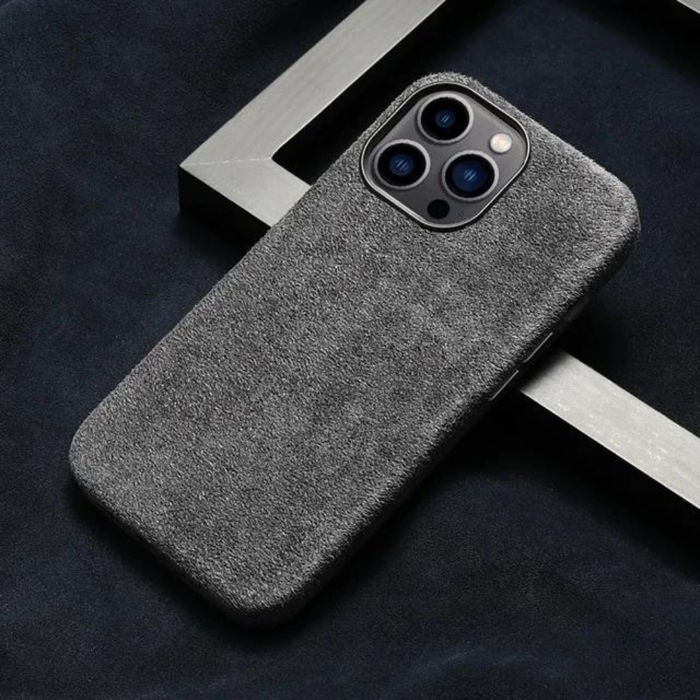 A smartphone with a textured gray suede finish case lies on a dark surface, partially framed by a metallic square object.