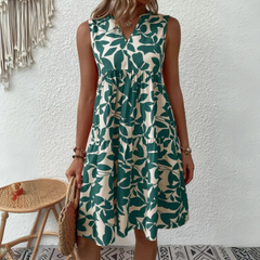 A woman stands against a white textured wall, wearing a sleeveless dress in a lightweight breathable fabric with a green leaf pattern—an ideal addition to your summer wardrobe. She stands by a wicker chair with a woven bag and macramé wall hanging nearby.