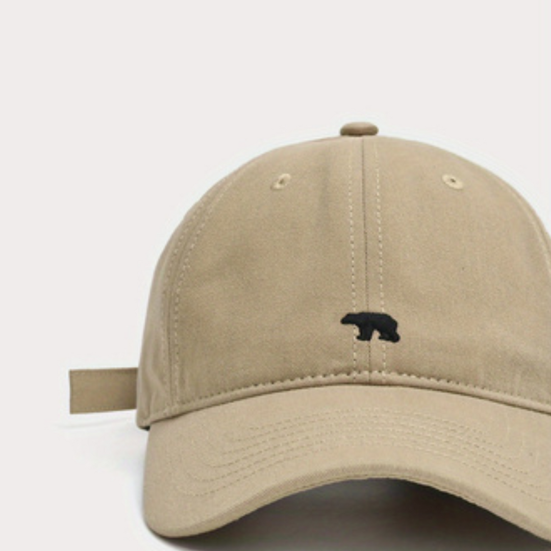A beige baseball cap crafted from premium cotton, featuring a small black bear embroidered on the front panel, shown against a plain white background.