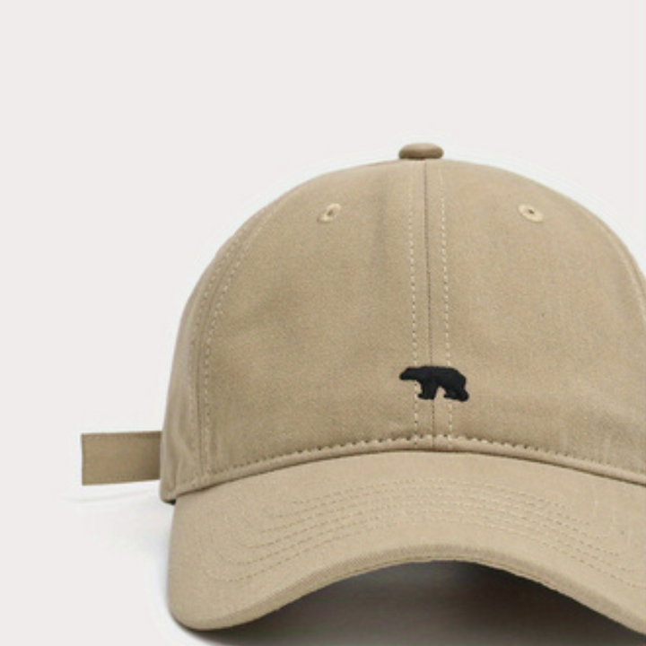 A beige baseball cap crafted from premium cotton, featuring a small black bear embroidered on the front panel, shown against a plain white background.