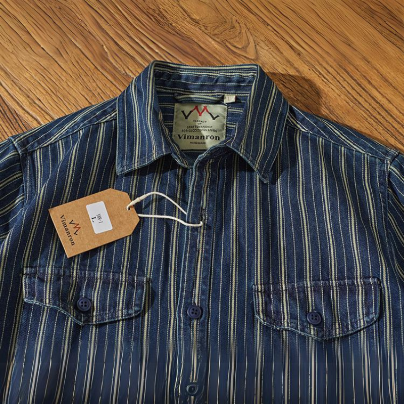 A relaxed-fit button-down blue denim shirt with yellow vertical stripes, two front pockets, and button closures is laid flat on a wooden surface. A brown price tag with the label Vimanron is attached to a buttonhole.