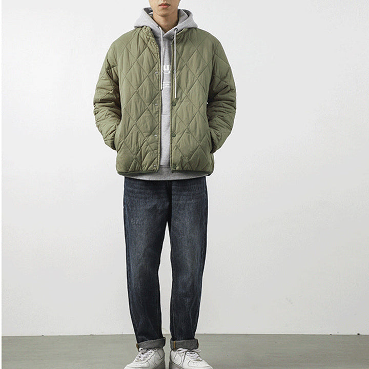 A person stands against a plain white wall, wearing an olive green padded jacket with a diamond-stitched pattern over a gray hoodie, dark jeans, and white sneakers. Their hands are in their jacket pockets, and their face is not fully visible.