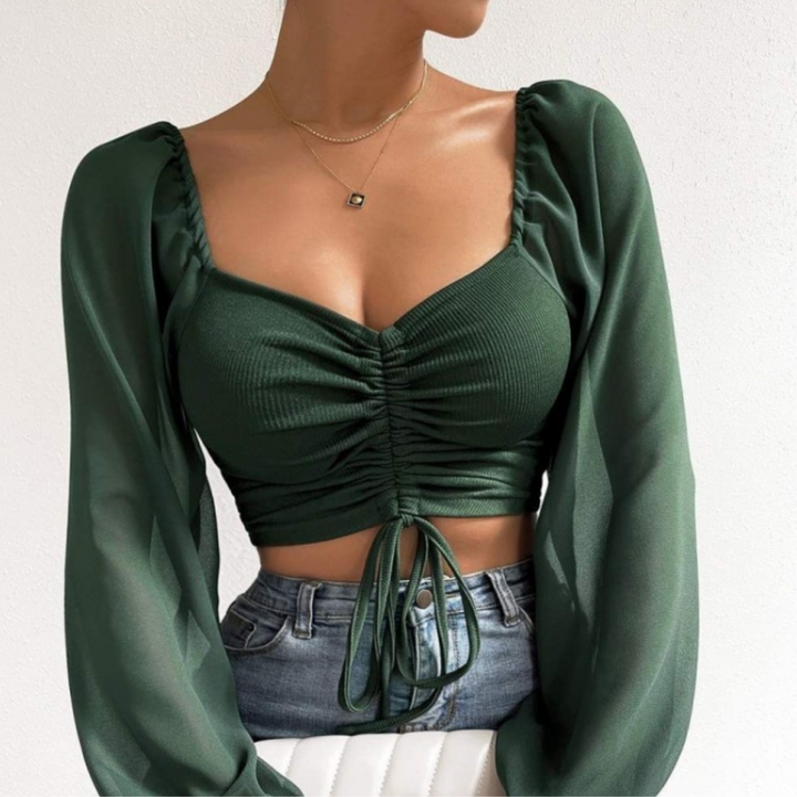 Woman wearing a dark green, long-sleeved crop top featuring a ruched front and tie at the waist, paired with light blue jeans and a simple gold necklace. She is seated against a plain white background.