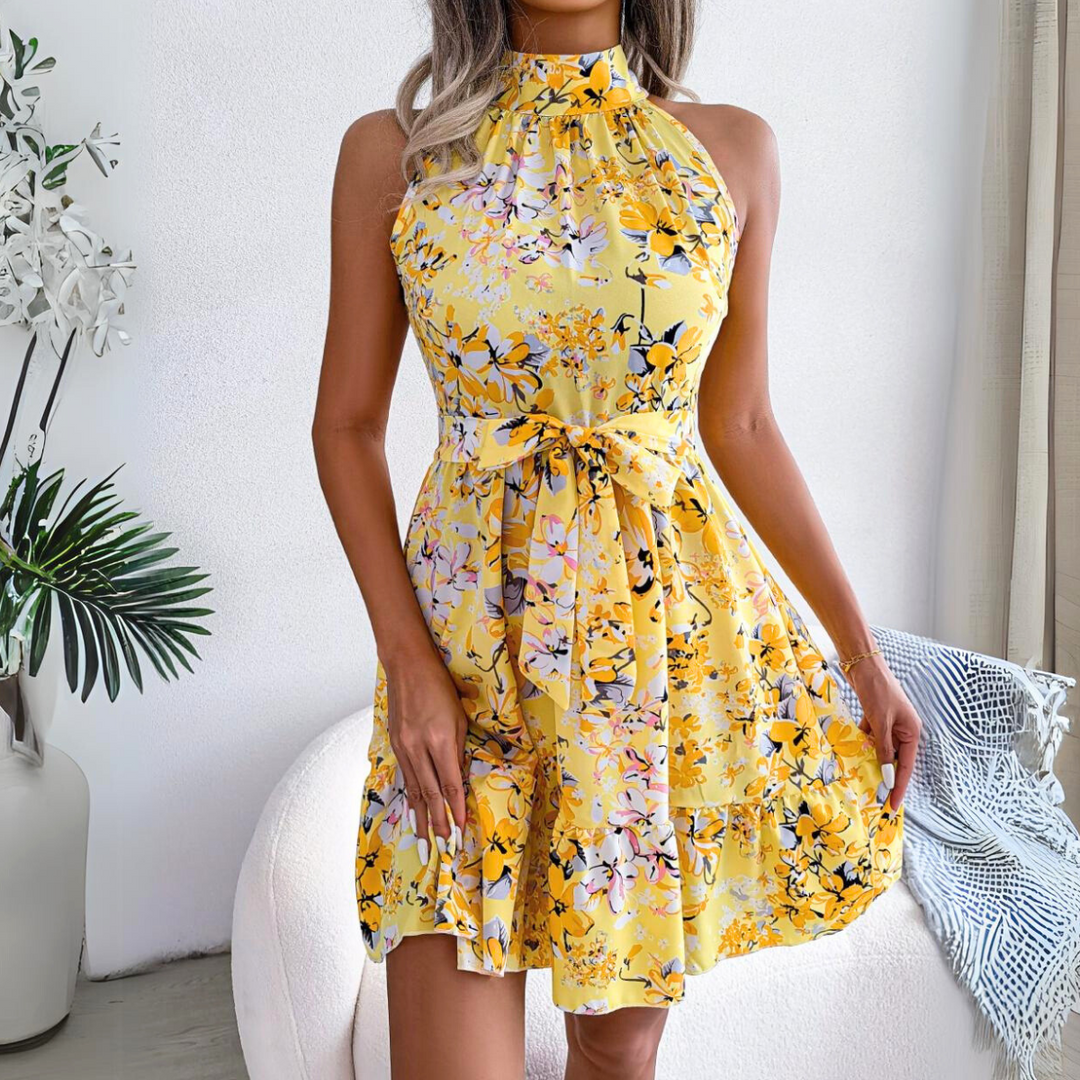 Woman wearing a sleeveless, halter neck yellow floral dress with a bow at the waist, standing indoors next to a white chair and green plants. A fresh pick for your summer wardrobe.