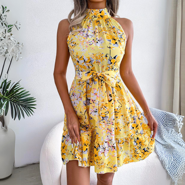 Woman wearing a sleeveless, halter neck yellow floral dress with a bow at the waist, standing indoors next to a white chair and green plants. A fresh pick for your summer wardrobe.