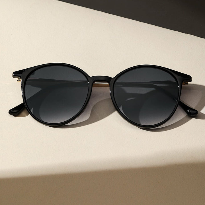 Round black sunglasses with dark lenses and UV protection rest on a light beige surface, with a shadow partially cast across the scene.