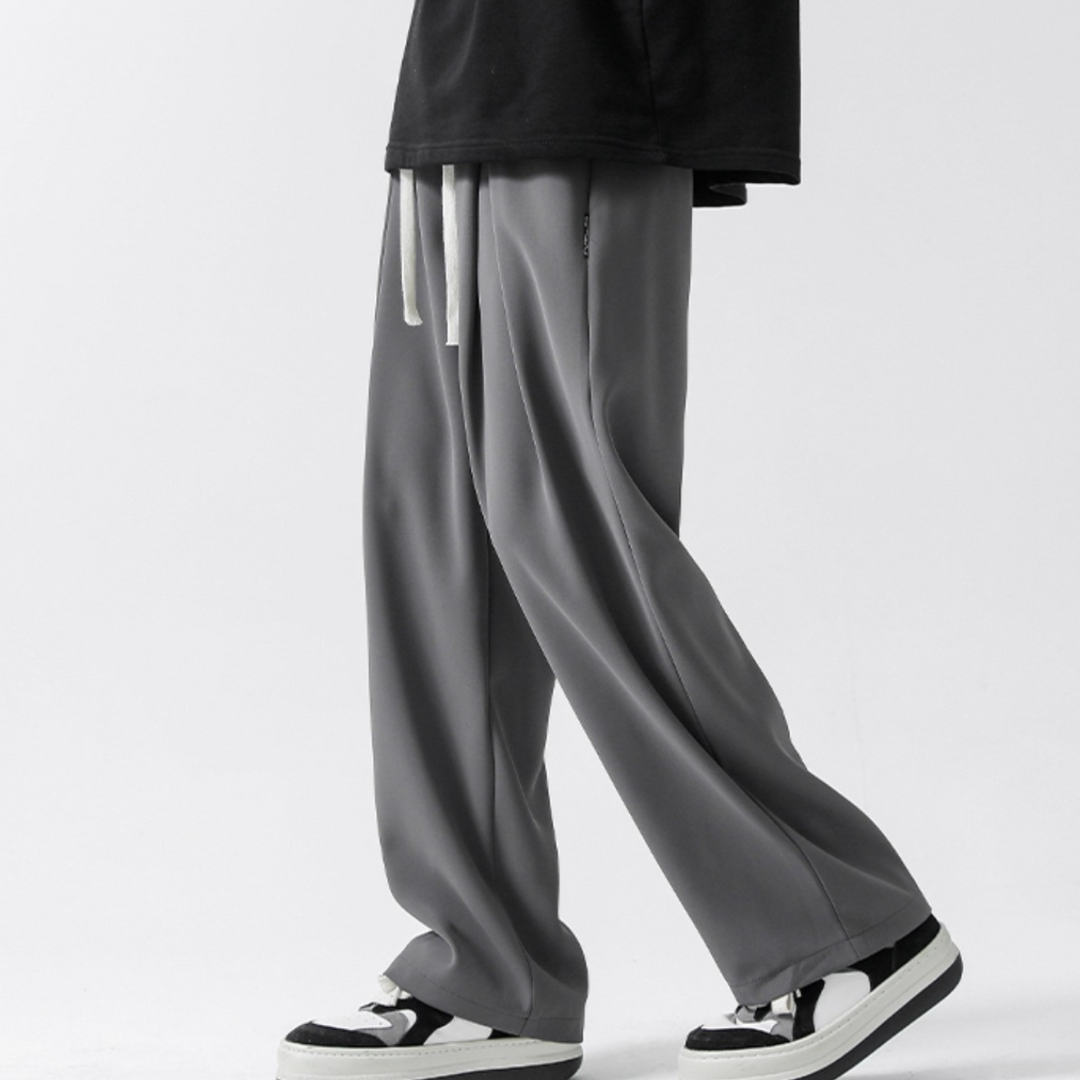 A person wearing loose-fitting gray streetwear joggers with white drawstrings, a black shirt, and black-and-white sneakers, standing against a plain white background.