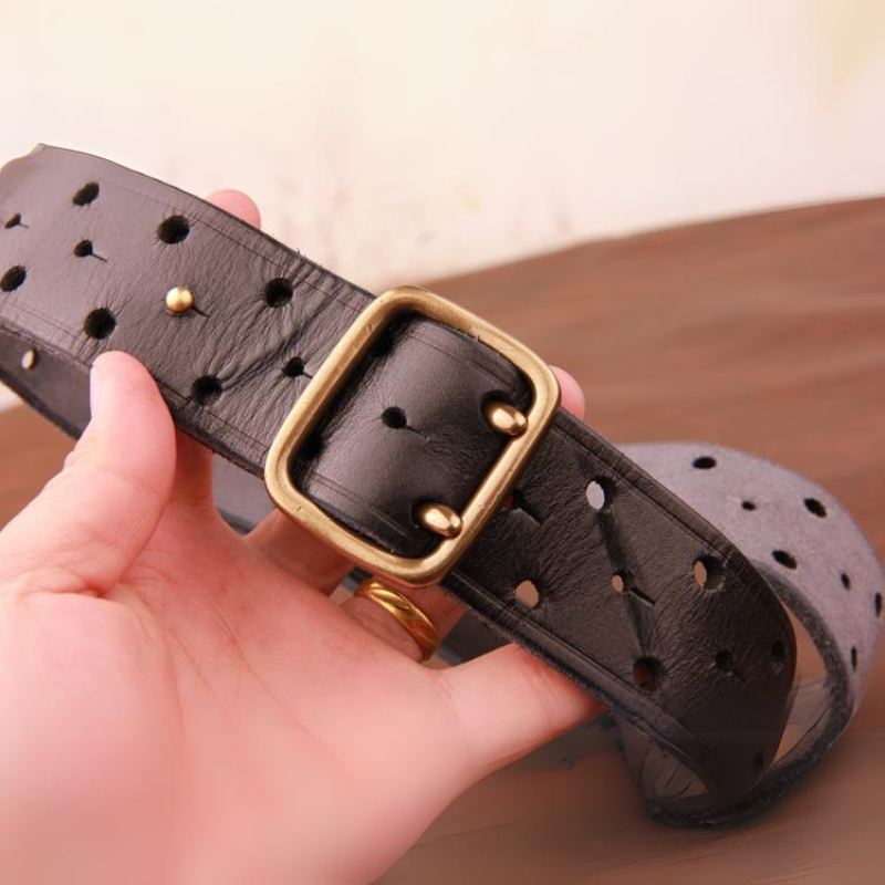 A hand holding a vintage leather belt with evenly spaced holes and a square, gold-colored metal buckle, against a blurred brown surface background.