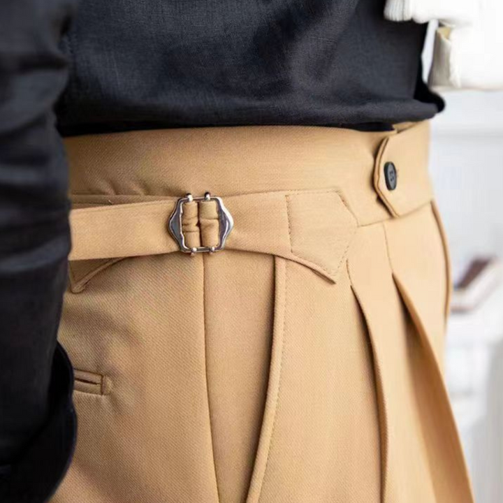 Close-up of a person wearing tailored fit tan pleated trousers with side adjusters and a black shirt tucked in. The metallic buckle and button detail on the waistband capture an old money aesthetic.