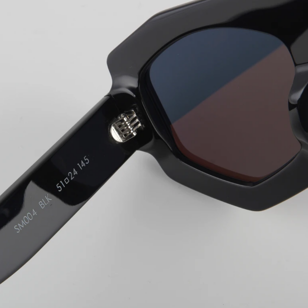 Close-up of black sunglasses featuring a geometric frame with dark lenses and UV400 protection. The hinge and acetate arm are shown, marked with text: SM004 BLK 51024 145 on a plain light gray background.