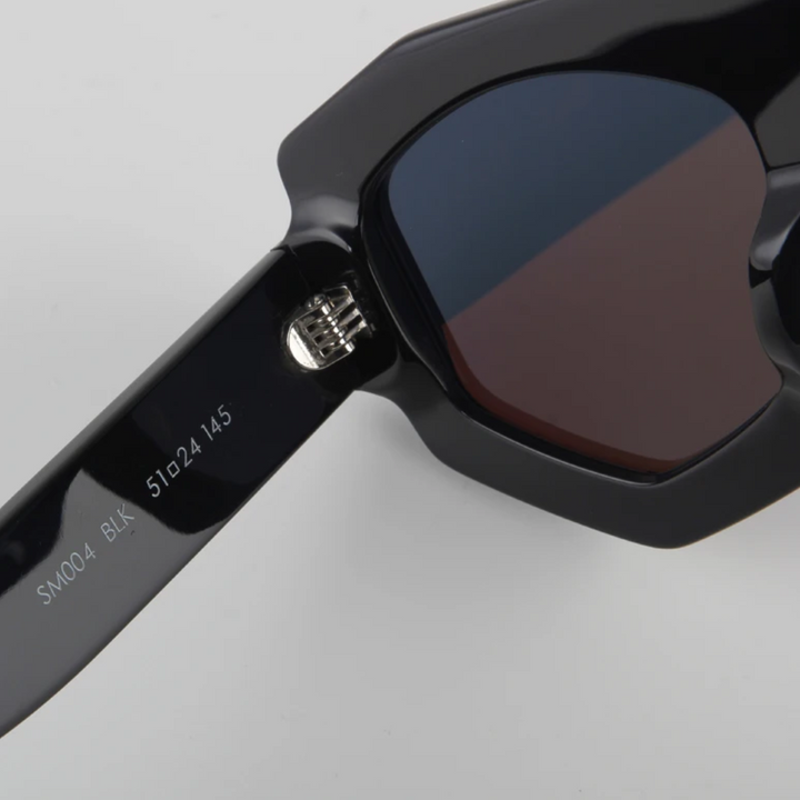 Close-up of black sunglasses featuring a geometric frame with dark lenses and UV400 protection. The hinge and acetate arm are shown, marked with text: SM004 BLK 51024 145 on a plain light gray background.
