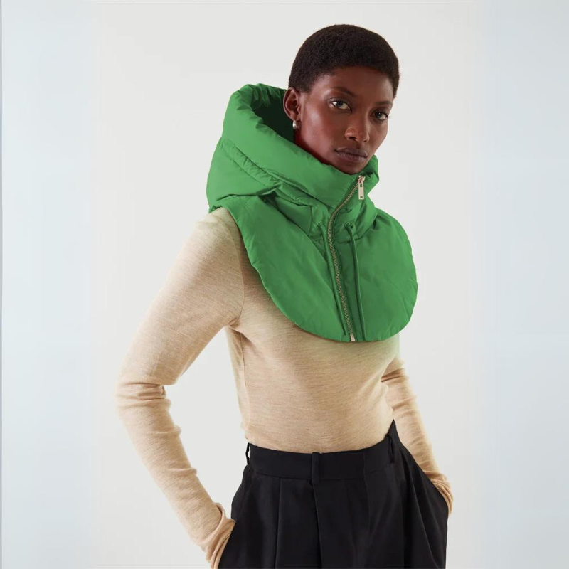 A person wearing a beige long-sleeve top, black trousers, and a lightweight puffy green hooded scarf with a zip-up front, standing against a plain white background.