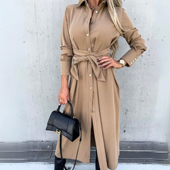 A woman wearing a long-sleeve wrap dress in tan with a waist tie, holding a black handbag and wearing a watch, stands against a concrete wall. Her long blonde hair is styled loosely, capturing effortless high fashion streetwear vibes.