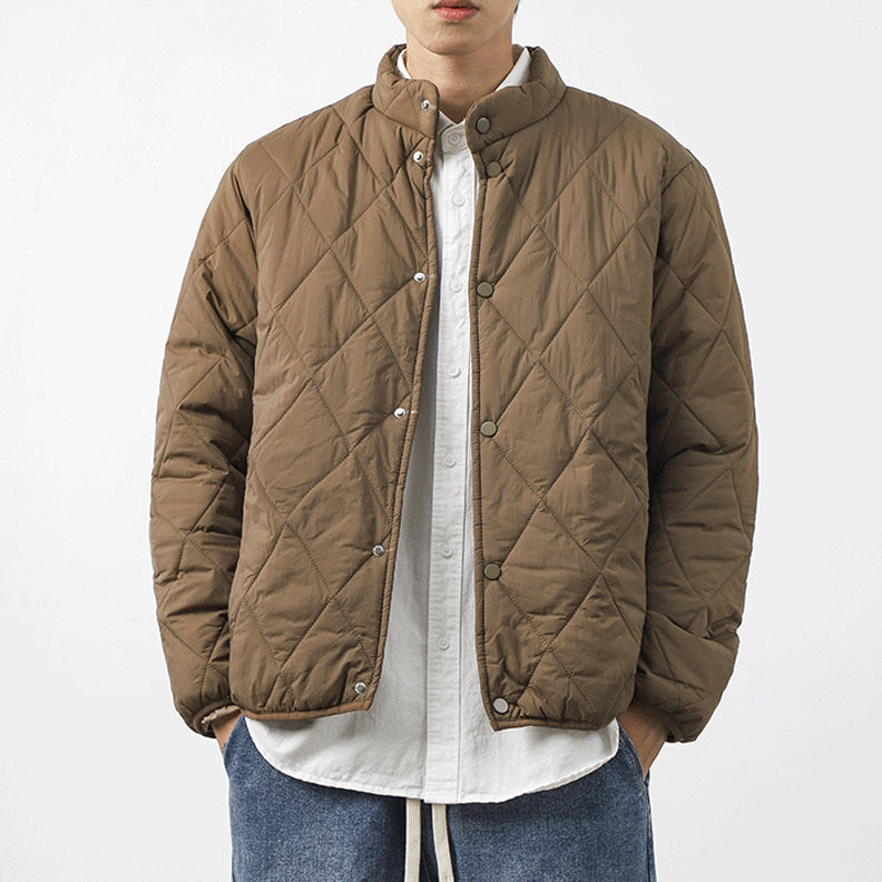 A person wearing a brown padded jacket with a diamond-stitched pattern over a white button-up shirt and blue jeans, standing against a plain white background. The lightweight insulating jacket features snap buttons and a stand collar. Face is partially out of frame.