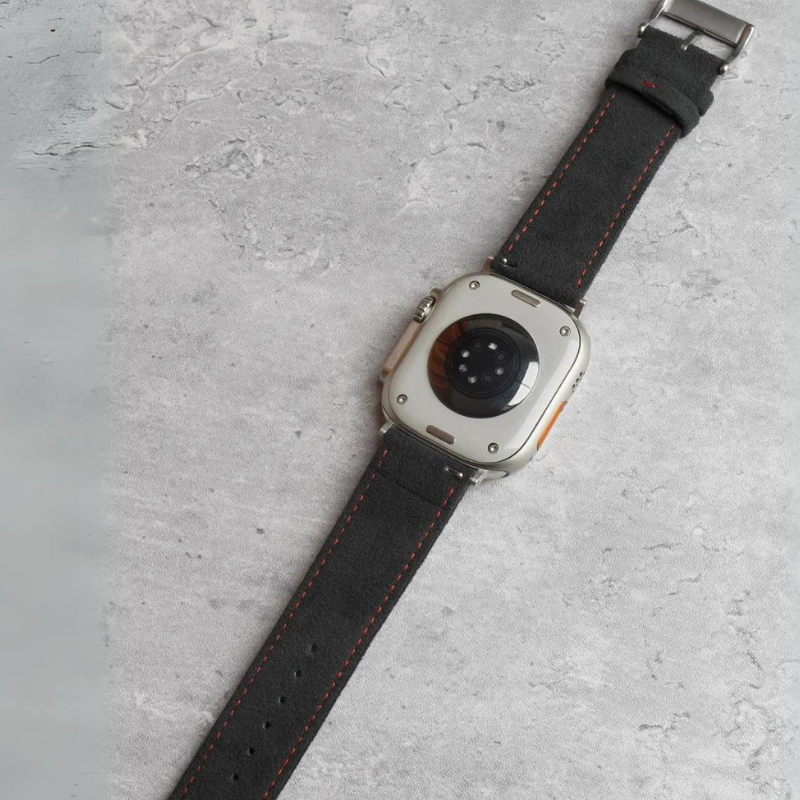 A smartwatch with a black leather strap and orange stitching, featuring a precision fit and stainless steel buckle, is shown face down on a light gray, textured surface. The watch’s sensors and metal casing are visible.