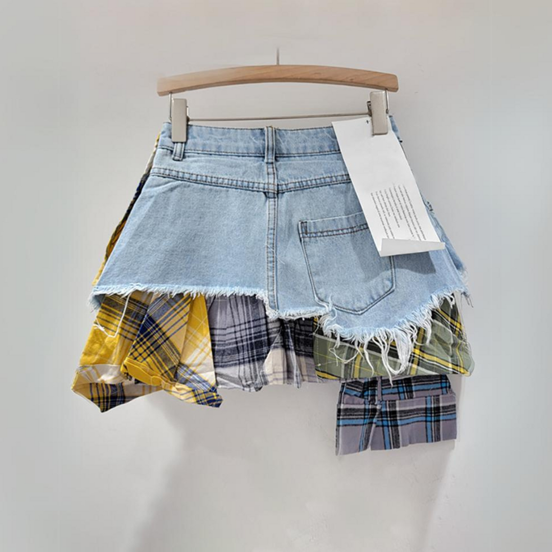 A short patchwork skirt with a frayed hem and eclectic denim and plaid panels in yellow, blue, and green, hanging on a wooden hanger against a plain light background.