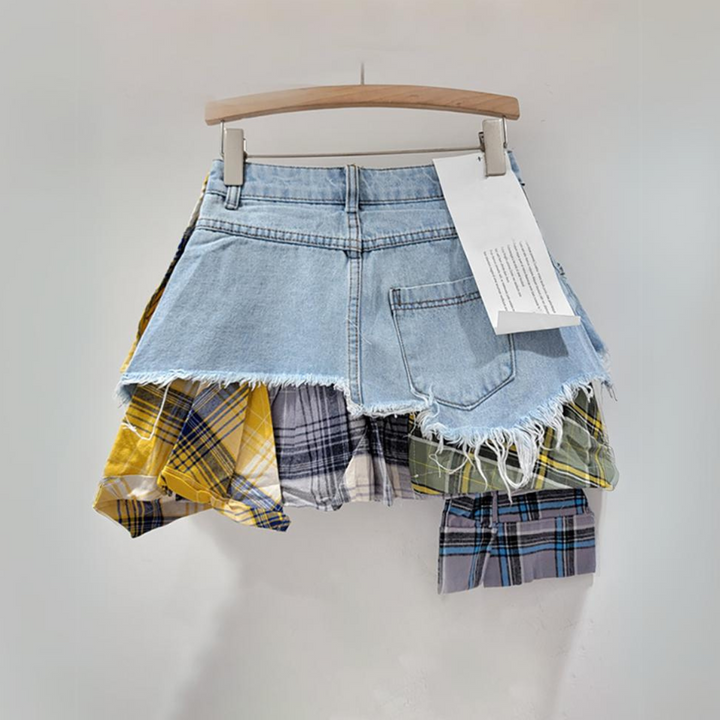 A short patchwork skirt with a frayed hem and eclectic denim and plaid panels in yellow, blue, and green, hanging on a wooden hanger against a plain light background.