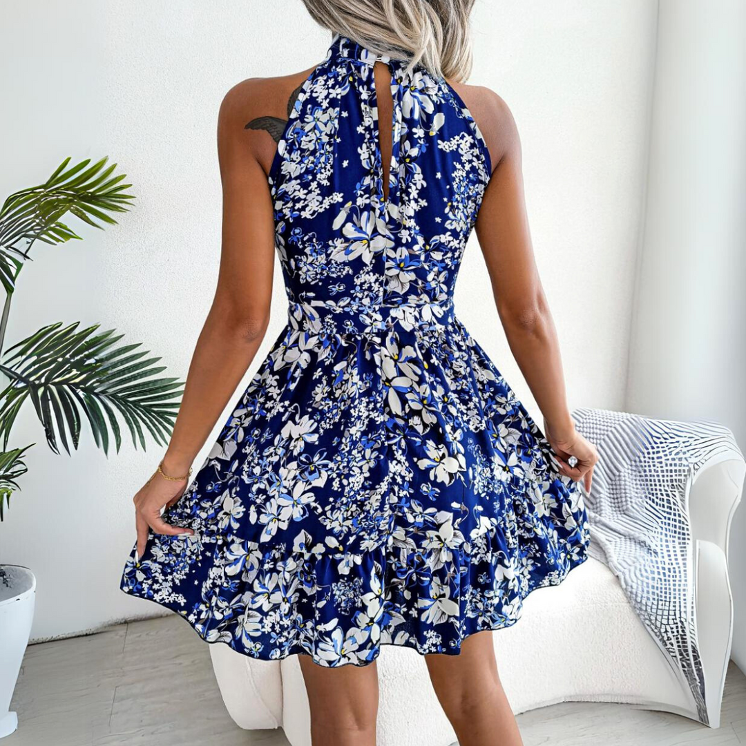 A woman wearing a sleeveless, halter neck blue and white floral dress poses with her back to the camera in a bright room with plants and a white armchair, perfect for updating your summer wardrobe.