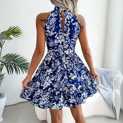 A woman wearing a sleeveless, halter neck blue and white floral dress poses with her back to the camera in a bright room with plants and a white armchair, perfect for updating your summer wardrobe.