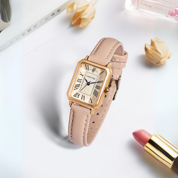 A gold rectangular face watch with a genuine leather strap rests on a white surface, surrounded by pink lipstick, perfume, and dried flowers. The watch features elegant Roman numerals and striking blue hands.