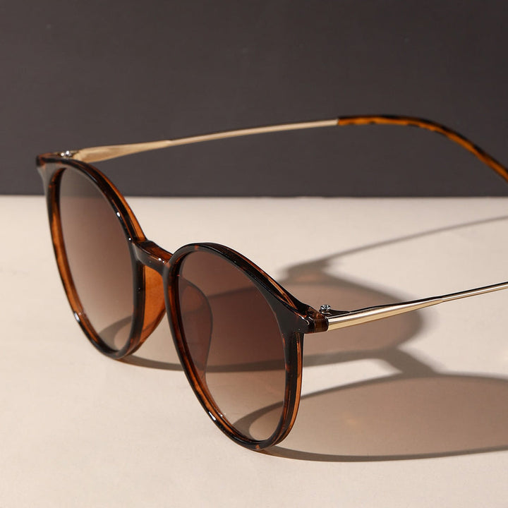 A pair of round, brown tortoiseshell sunglasses with gold metal arms rests on a light surface with a dark background, offering UV protection and anti-glare features.