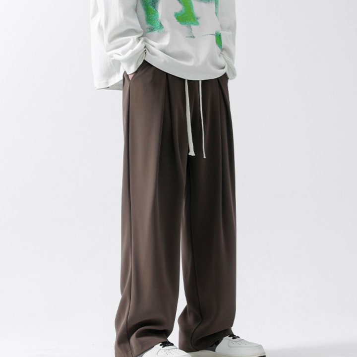 Person wearing loose, dark brown streetwear joggers with a drawstring and a white long-sleeve shirt featuring green designs, hands in pockets, and white sneakers, standing against a plain white background.