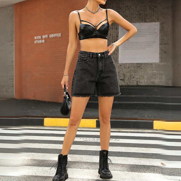A woman stands on a crosswalk wearing a black leather crop top, black wash high-waist denim shorts, combat boots, a necklace, and holds a small black handbag, with a brick building in the background.