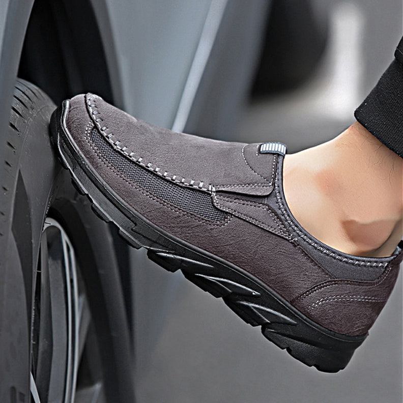 A close-up of a person's foot wearing gray breathable loafers, pressing against a vehicle tire. The shoe has a textured sole, visible stitching along the upper part, and offers a comfortable fit for everyday wear.