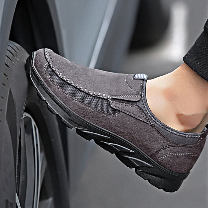 A close-up of a person's foot wearing gray breathable loafers, pressing against a vehicle tire. The shoe has a textured sole, visible stitching along the upper part, and offers a comfortable fit for everyday wear.