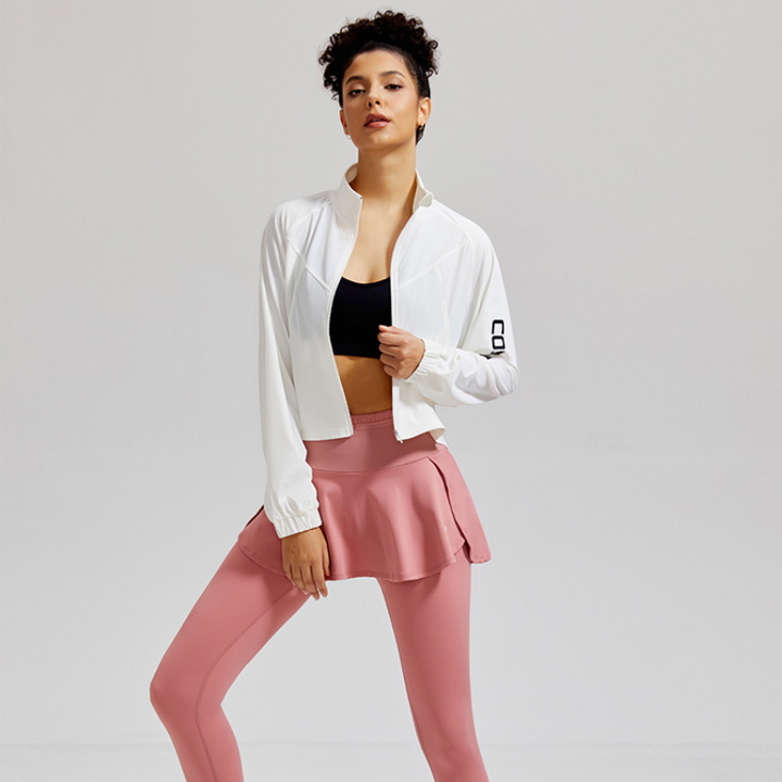 A woman poses confidently in athletic wear, featuring high-waisted support skirted leggings with stretch storage pockets, a white zip-up jacket, and a black sports bra, standing against a light gray background.