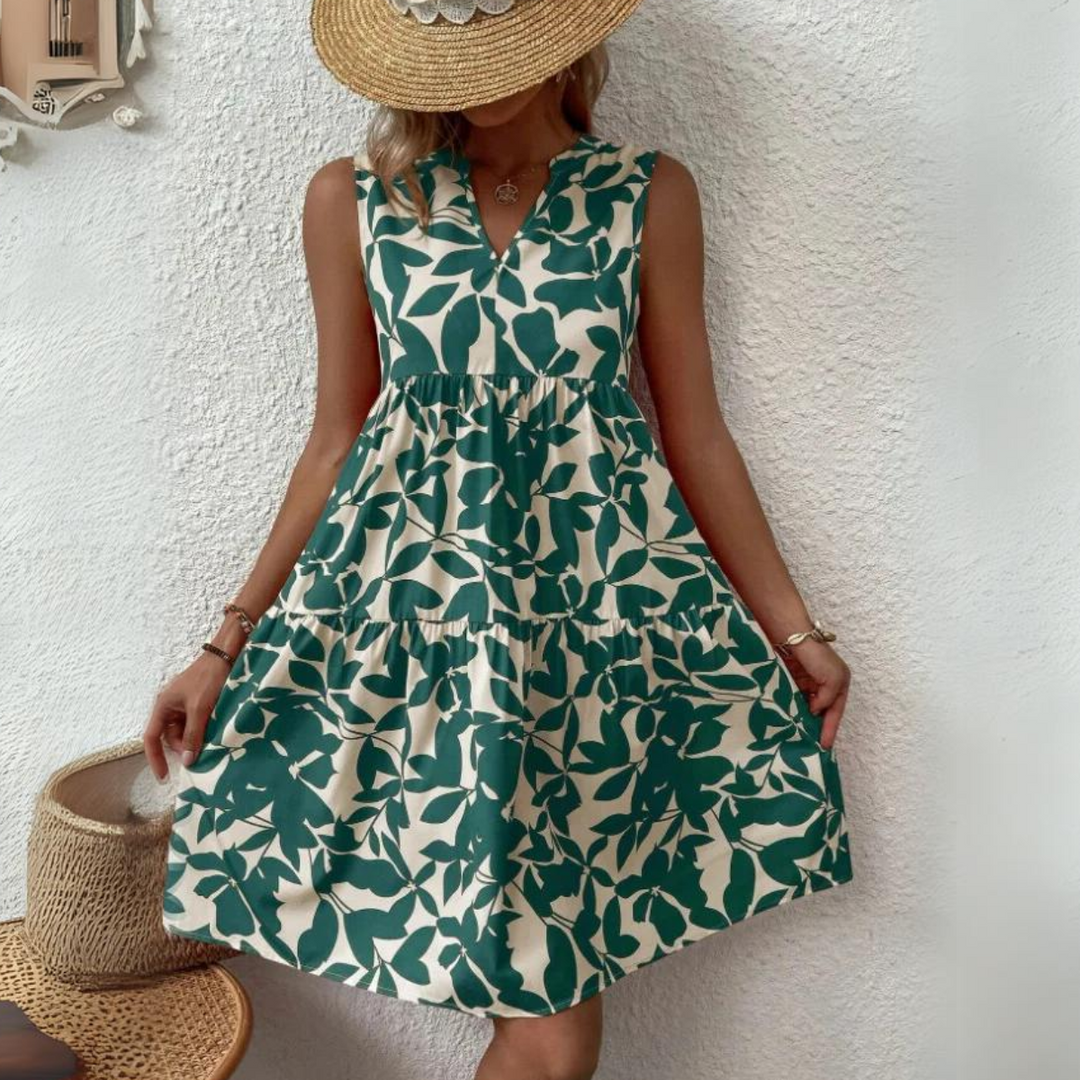 A woman in a sleeveless dress made from lightweight breathable fabric, featuring a green leaf pattern, stands against a white textured wall. She holds the sides of her dress—an elegant staple for any summer wardrobe—wearing a wide-brimmed straw hat.