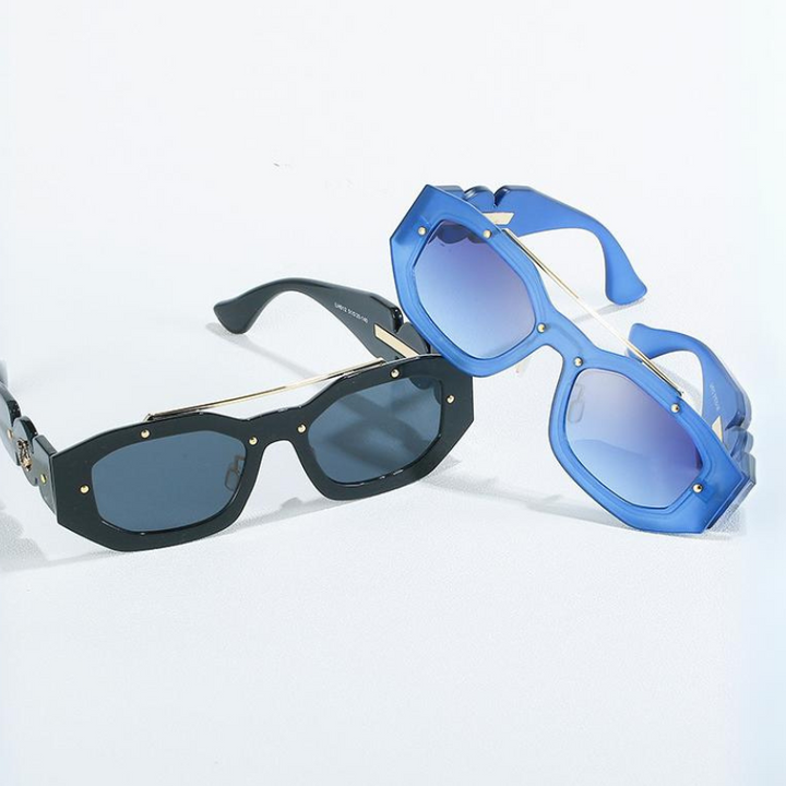 Two pairs of sunglasses on a white surface, both featuring scratch-resistant lenses and gold accents: one with black rectangular frames and dark lenses, the other with blue geometric frames and blue-tinted lenses.